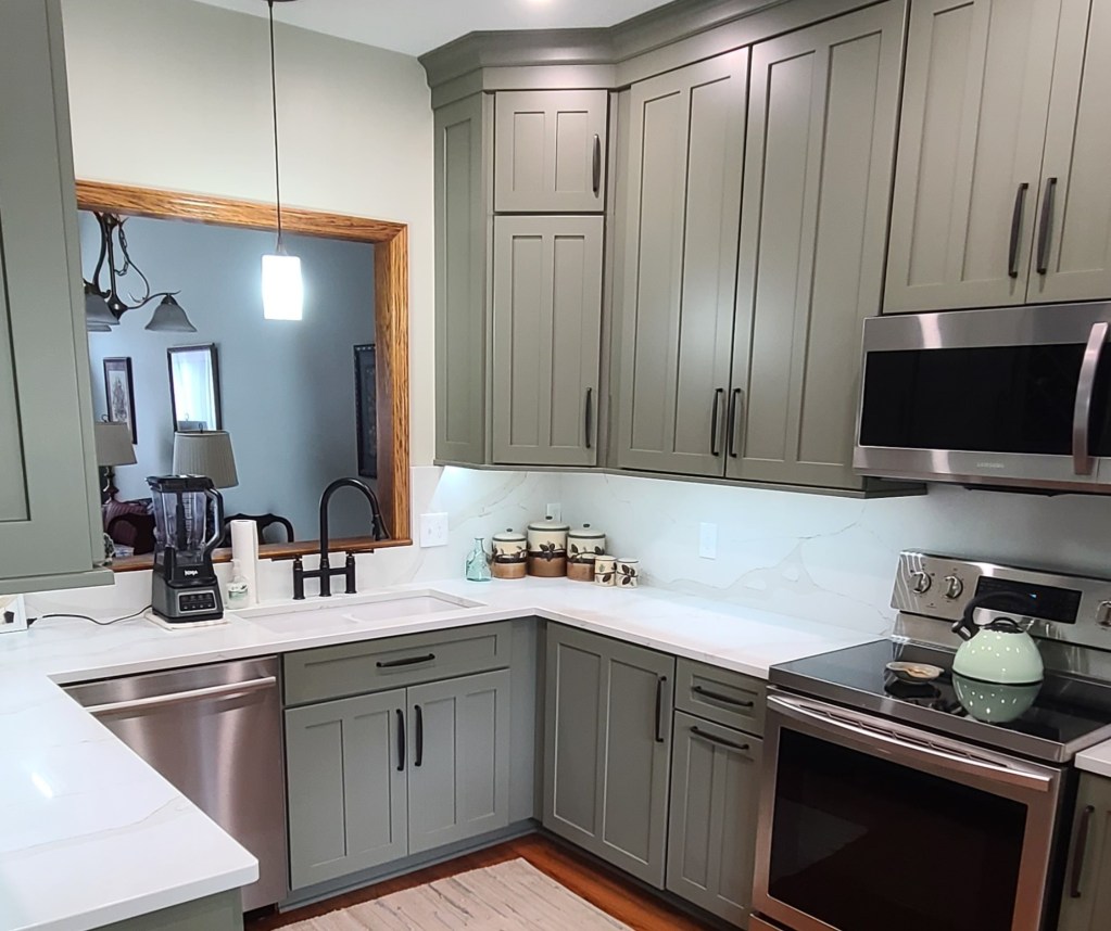 A remodeled kitchen with green shaker-style cabinets, white quartz countertops, and stainless steel appliances. The kitchen features hardwood floors, recessed lighting, and a large sink with a black faucet. A wooden-framed pass-through window connects the kitchen to the adjacent room.