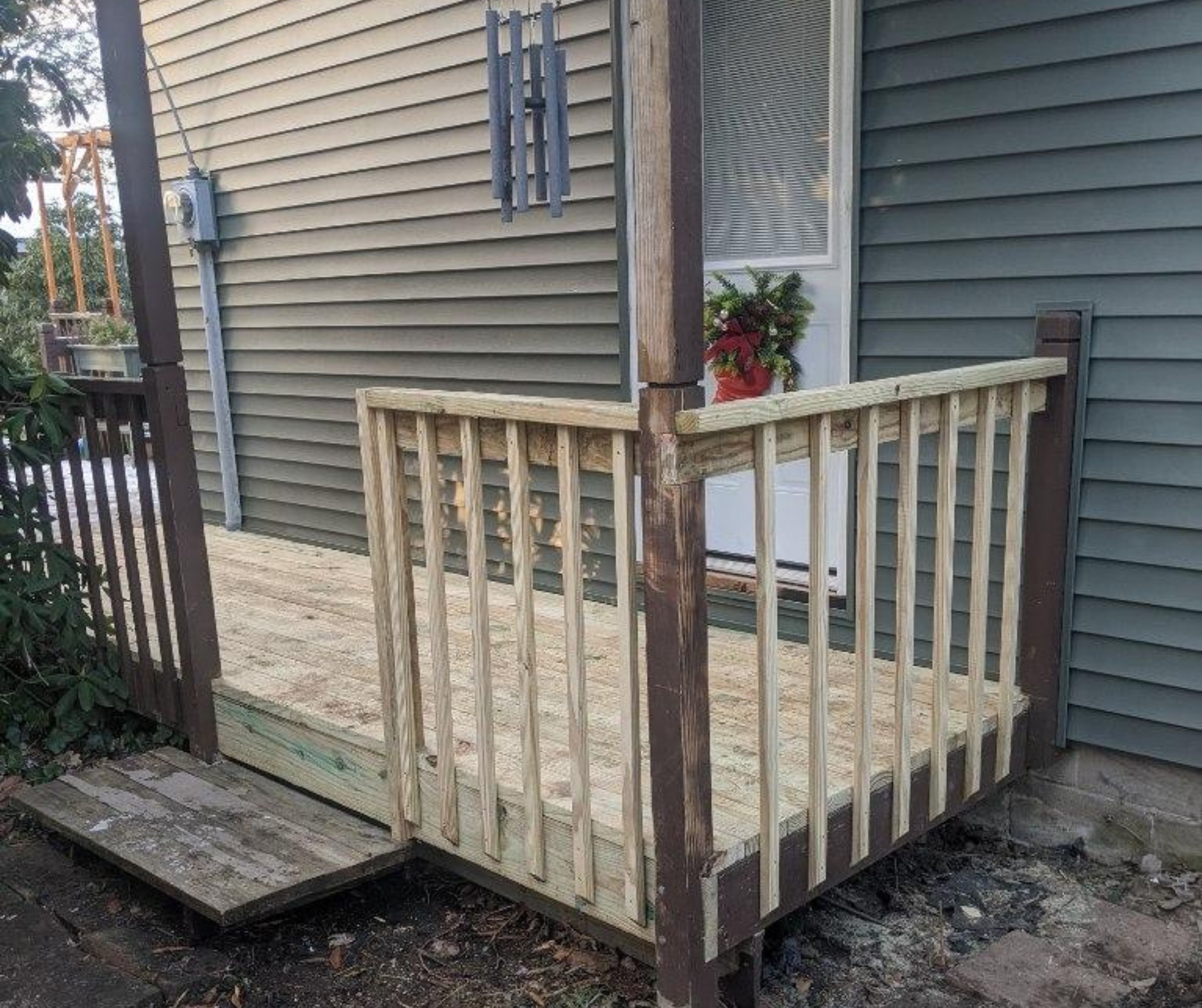 A newly constructed wooden porch with railing, attached to a home with gray siding. The porch features simple, clean lines with natural wood tones and a festive wreath on the door. A wind chime hangs from the porch roof, adding a decorative touch to the space.