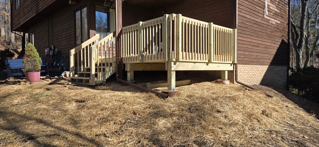 A newly constructed wooden deck with stairs, railings, and support posts attached to a brown house on a sloped yard covered in straw mulch. The deck remodel is completed with pressure-treated lumber.