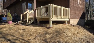 A newly constructed wooden deck with stairs, railings, and support posts attached to a brown house on a sloped yard covered in straw mulch. The deck remodel is completed with pressure-treated lumber.