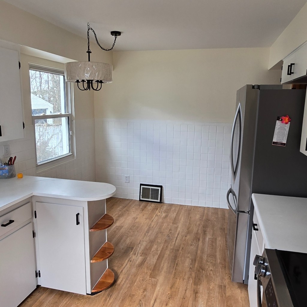 Updated kitchen featuring white cabinets, wood-look flooring, stainless-steel refrigerator, white countertops, black hardware, and a new modern chandelier above an open dining nook.