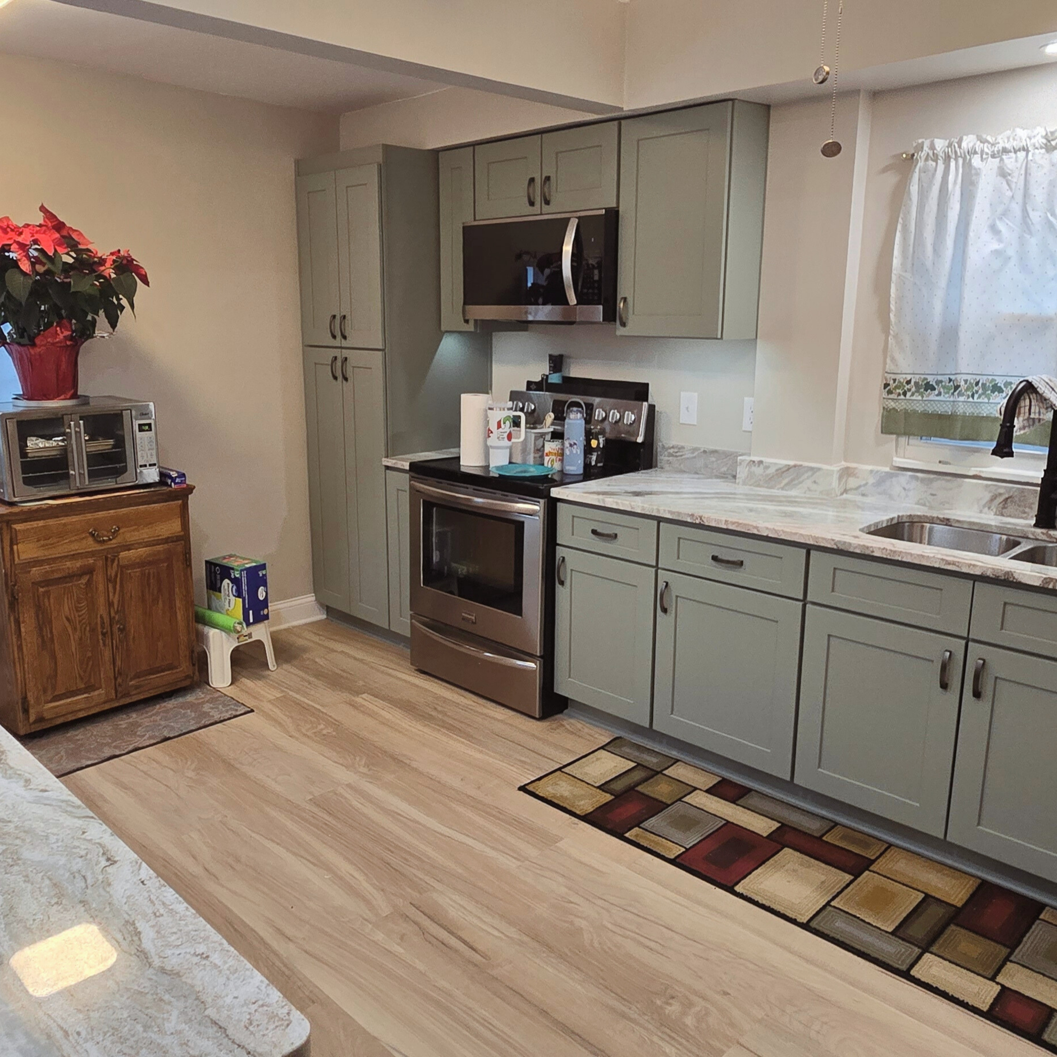 Wide view of a remodeled kitchen featuring sage cabinetry, new appliances, luxury vinyl plank flooring, and updated countertops.