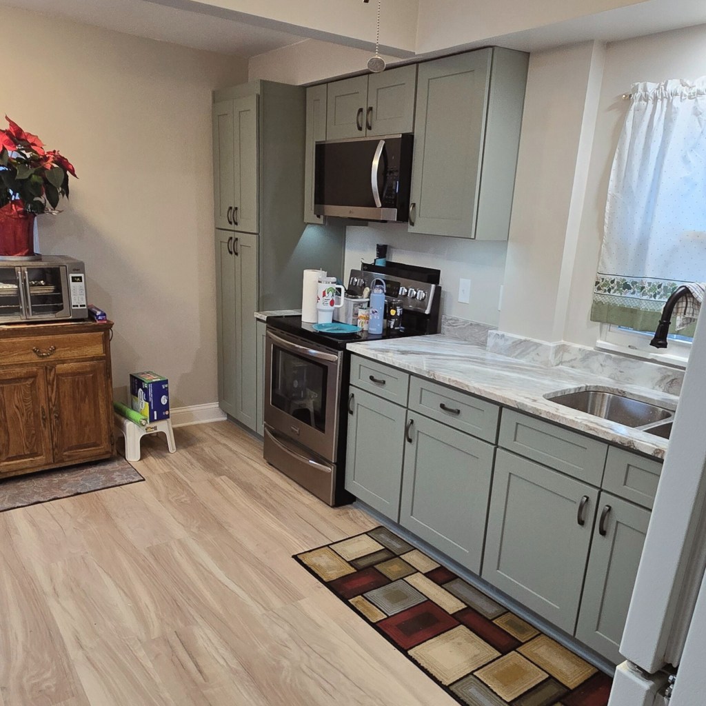 Kitchen showing stainless range and microwave, sage green cabinets, marble-look countertops, and light wood-look flooring.
