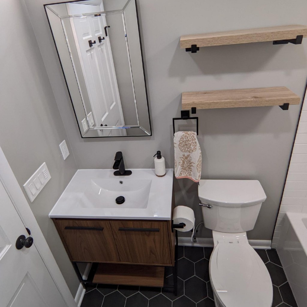 Updated bathroom vanity with wood cabinetry, white countertop, black faucet, modern mirror, and hex tile flooring completed by local remodeling contractors.