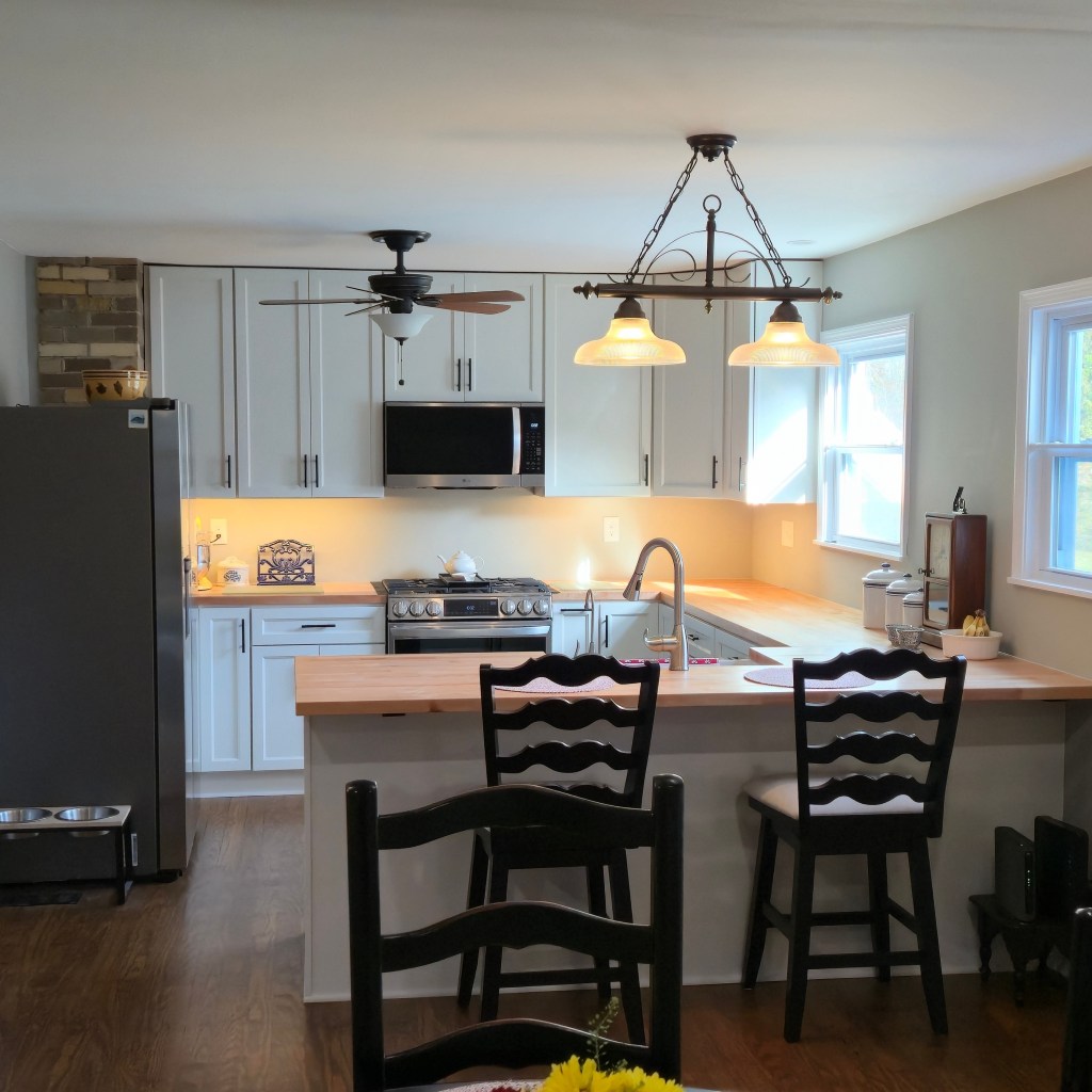 Remodeled kitchen featuring white shaker cabinets, butcher block countertops, stainless steel appliances, breakfast bar seating, new lighting, and updated flooring.