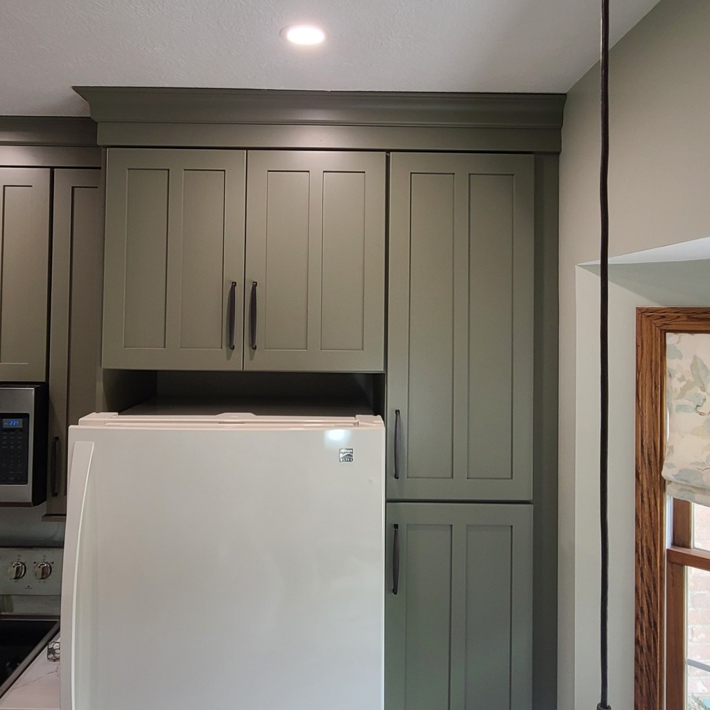 Tall sage green kitchen cabinets surrounding a refrigerator, featuring shaker doors, modern hardware, and crown molding.