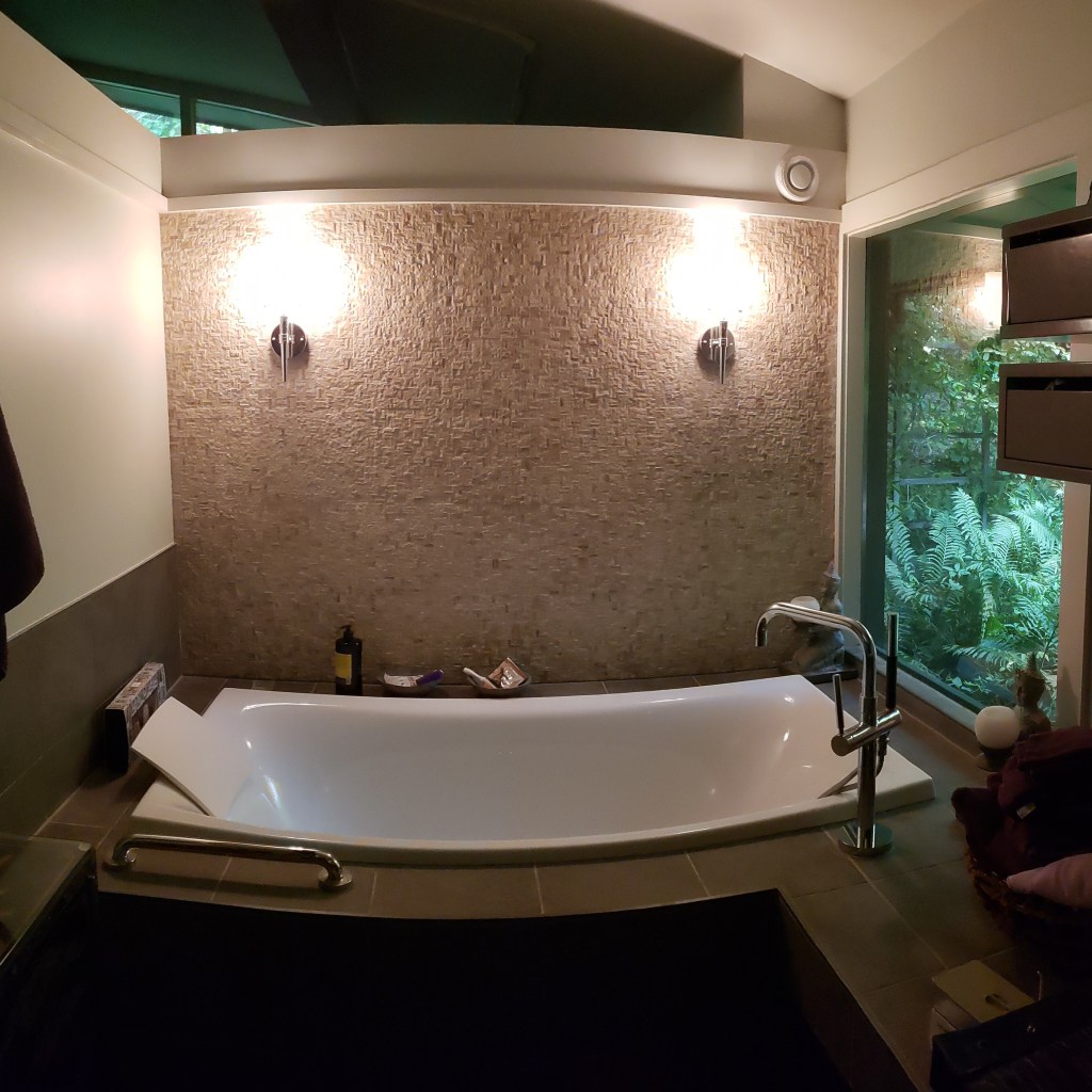 Freestanding soaking tub with textured tile accent wall and modern wall sconces in a luxury bathroom remodel