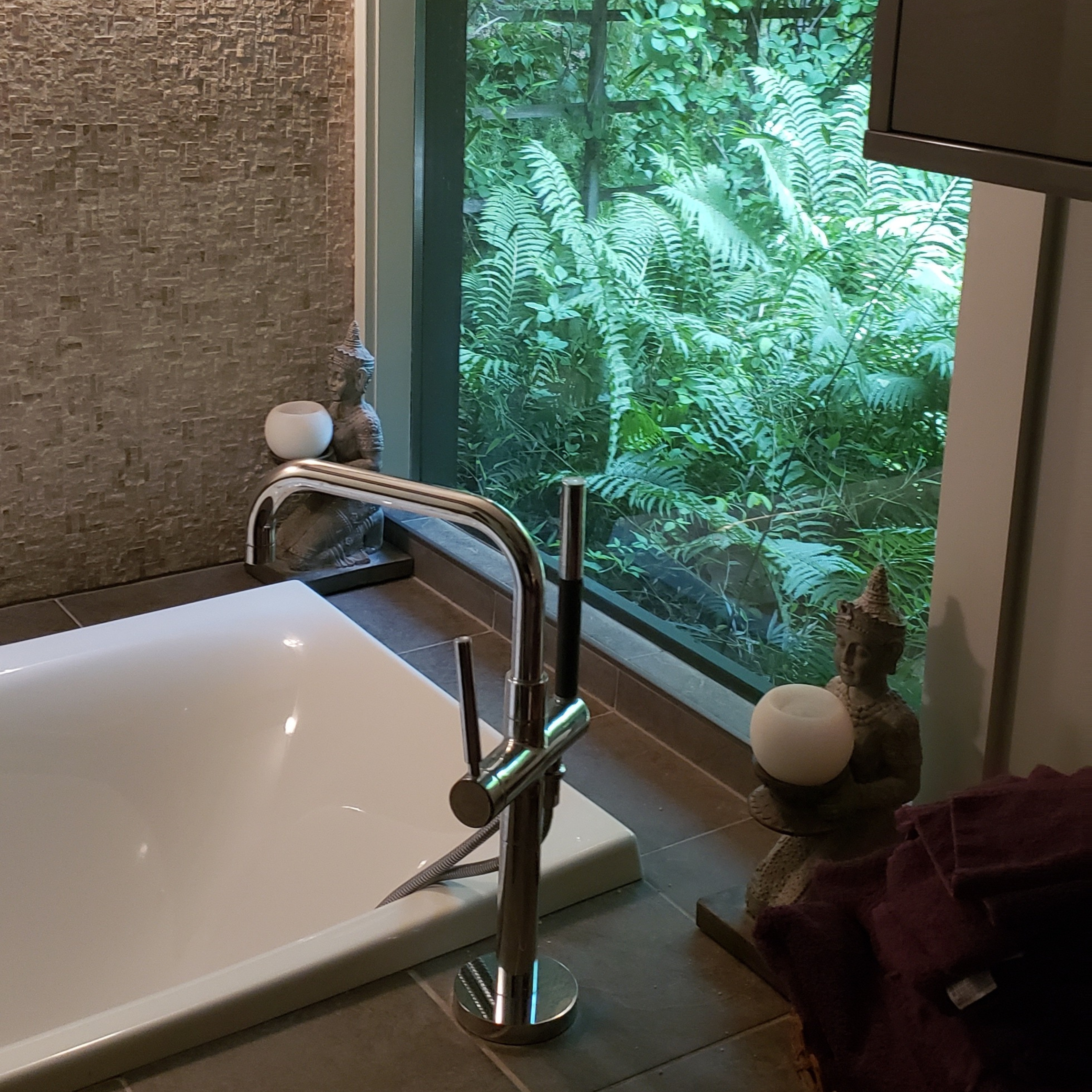 Floor-mounted tub faucet next to freestanding bathtub with large window overlooking greenery