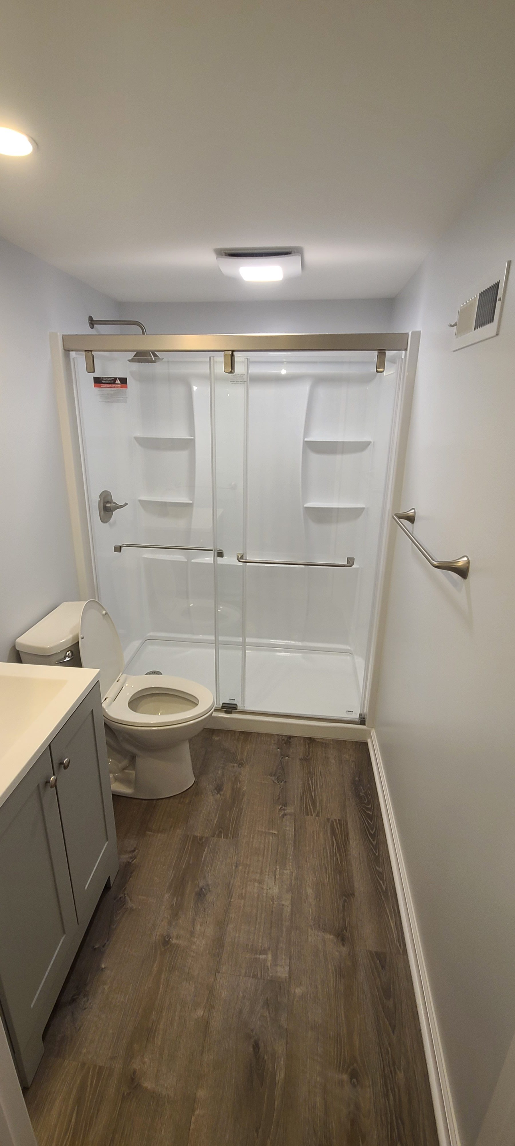 Bathroom remodel in Akron, Ohio featuring a walk-in shower with glass sliding doors, gray vanity, white countertop, brushed nickel fixtures, grab bars, and wood-look flooring.